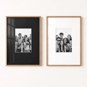 May include: Two rectangular wooden frames with light brown edges. The left frame features a black mount surrounding a black and white photograph of a group of friends. The right frame features a white mount surrounding a black and white group photo.