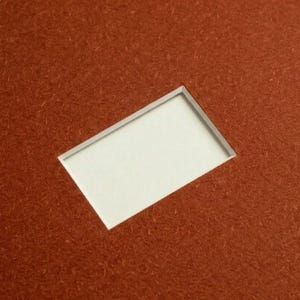 May include: A rectangular piece of textured, rust-colored material featuring a clean, centered rectangular cutout. The interior of the cutout reveals a smooth, plain white surface, creating a sharp contrast between the deep orange-brown exterior and the bright white center.