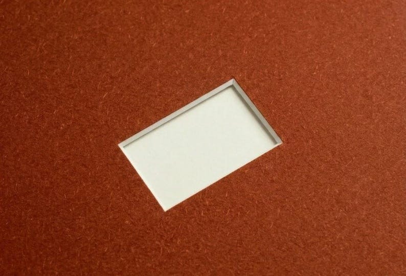 May include: A rectangular piece of textured, rust-colored material featuring a clean, centered rectangular cutout. The interior of the cutout reveals a smooth, plain white surface, creating a sharp contrast between the deep orange-brown exterior and the bright white center.