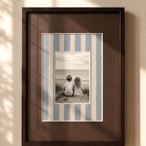 Könnte beinhalten: Ein rechteckiger Bilderrahmen aus dunkelbraunem Holz mit einem Schwarz-Weiß-Foto von zwei Kindern, die auf einer grasbewachsenen Düne sitzen und auf das Meer blicken. Das Foto ist von einem Passepartout mit hellblauen und cremefarbenen Streifen umgeben.