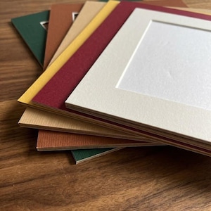 Pode incluir: Uma pilha de passe-partouts retangulares de papel com uma variedade de cores, incluindo creme, vermelho escuro, amarelo mostarda, castanho claro, terracota e verde escuro. Cada passe-partout tem um recorte retangular central com borda chanfrada.