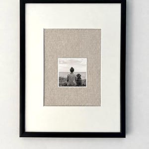 Könnte beinhalten: Ein Schwarz-Weiß-Foto in einem schwarzen Rahmen mit einem breiten weißen Passepartout und einem inneren Rand aus strukturiertem beigem Leinen. Das Bild zeigt eine Person und einen Hund, die nebeneinander sitzen und von hinten auf das Wasser blicken.