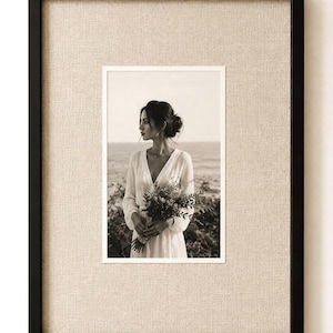 Pode incluir: Uma moldura retangular preta contendo uma fotografia em tom sépia de uma pessoa com o cabelo num coque, vestindo um vestido de manga comprida e segurando um ramo de flores. A foto é rodeada por um passe-partout de tecido bege claro texturado.