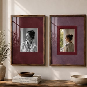 May include: Two rectangular wall frames with dark wood borders and deep burgundy matting. Each frame contains a black and white portrait of a person looking out a window. The frames are displayed side by side on a wall above a wooden surface.