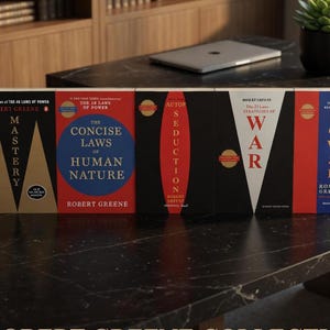 May include: A collection of Robert Greene books, including "Mastery", "The Concise Laws of Human Nature", "Seduction", and "The 33 Strategies of War". The books are on a black marble surface with a laptop and a small plant.