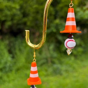 K&ouml;nnte beinhalten: Ein Paar ausgefallene Ohrringe mit orange-wei&szlig; gestreiften Verkehrskegeln, Baseb&auml;llen und goldfarbenen Haken. Die Ohrringe h&auml;ngen an einem goldfarbenen, gebogenen B&uuml;gel vor einem verschwommenen gr&uuml;nen Hintergrund.