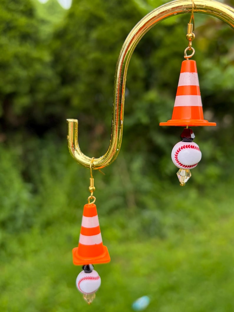 K&ouml;nnte beinhalten: Ein Paar ausgefallene Ohrringe mit orange-wei&szlig; gestreiften Verkehrskegeln, Baseb&auml;llen und goldfarbenen Haken. Die Ohrringe h&auml;ngen an einem goldfarbenen, gebogenen B&uuml;gel vor einem verschwommenen gr&uuml;nen Hintergrund.