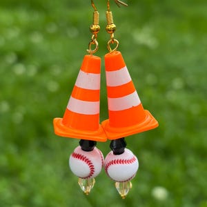 K&ouml;nnte beinhalten: Ein Paar ausgefallene Ohrringe mit orange-wei&szlig; gestreiften Verkehrskegel-Anh&auml;ngern, kombiniert mit Baseball-Themen-Perlen. Die Ohrringe haben goldfarbene Haken. Der Hintergrund ist verschwommen gr&uuml;n.