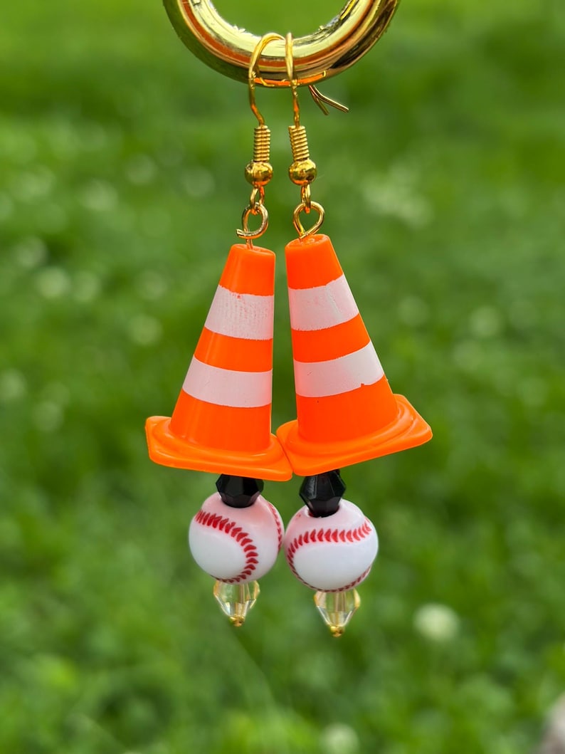 K&ouml;nnte beinhalten: Ein Paar ausgefallene Ohrringe mit orange-wei&szlig; gestreiften Verkehrskegel-Anh&auml;ngern, kombiniert mit Baseball-Themen-Perlen. Die Ohrringe haben goldfarbene Haken. Der Hintergrund ist verschwommen gr&uuml;n.
