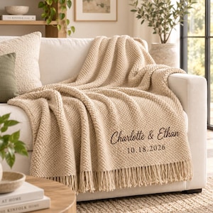 Op de afbeelding: Een beige gebreide plaid met een textuurpatroon en franjes. De deken is voorzien van donkerbruin geborduurde tekst met de namen Charlotte & Ethan en de datum 18-10-2026, gedrapeerd over een witte bank met een groen sierkussen.