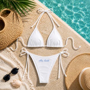 May include: A white bikini set featuring a triangle top with halter neck ties and matching side-tie bottoms. The bikini bottoms are embroidered with the text "Mrs. Smith" in light blue script lettering on the front centre of the fabric.