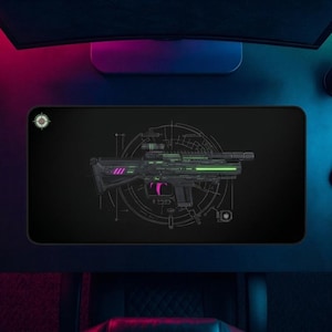 May include: A rectangular black desk mat featuring a central graphic of a futuristic rifle in shades of green and magenta. The design includes technical blueprint-style circular lines and markings, with a small circular emblem in the top left corner.
