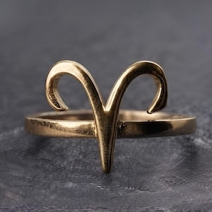 Könnte beinhalten: Ein goldfarbener Metallring mit dem Widder-Sternzeichen als zentralem Element. Das Design besteht aus einer glatten, kreisförmigen Ringschiene, die mit dem stilisierten Widderhorn-Motiv verbunden ist, das von der Vorderseite des Rings nach oben ragt.