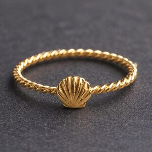 Puede incluir: Un anillo de color dorado con una banda fina de estilo cuerda trenzada y un pequeño dije de concha marina centrado. El metal tiene un acabado pulido y reflectante que resalta las crestas detalladas del diseño de la concha en la banda.