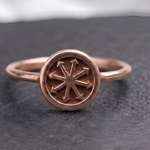 Op de afbeelding: Een roségouden ring met een rond vlak voorzien van een verhoogd achtpuntig ster-symbool. De ringband is dun en glad, en het centrale embleem toont een symmetrisch ontwerp met pijlen die vanuit het midden van de cirkel naar buiten wijzen.