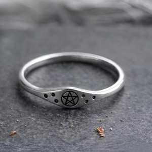 May include: A thin silver ring featuring a central circular emblem engraved with a five-pointed star inside a circle. The band has a slightly raised, tapered front section decorated with three small, dark, recessed dots on each side of the star.
