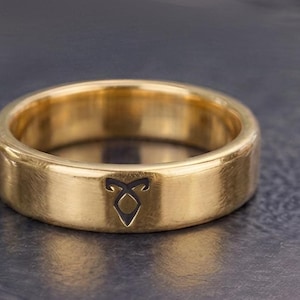 Könnte beinhalten: Ein polierter goldfarbener Bandring mit einer glatten, abgerundeten Außenfläche. Ein kleines, schwarzes, eingraviertes Symbol, das einem stilisierten Buchstaben A mit einer spitzen Oberseite und einer rautenförmigen Basis ähnelt, ist mittig.