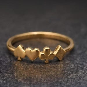 May include: A gold ring featuring four distinct card suit symbols arranged in a row along the top of the band. The design includes a spade, a heart, a club, and a diamond, all rendered in a polished gold finish with a smooth, rounded metal band.