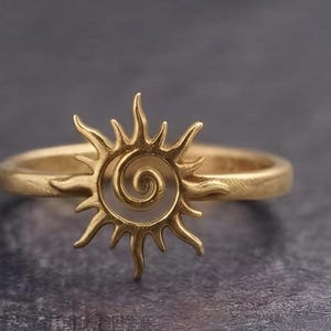 May include: A gold-toned ring featuring a central sun motif with a spiral centre and radiating pointed rays. The band has a textured, matte finish, and the sun design is integrated directly into the circular band structure of the jewellery piece.