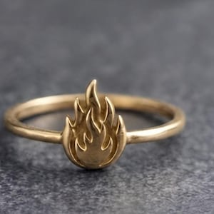 Può includere: Un anello color oro caratterizzato da un emblema centrale a forma di fiamma. La fascia è liscia e circolare, mentre il punto focale mostra un design a fuoco stilizzato e in rilievo con diverse lingue di fiamma appuntite rivolte verso l'alto.