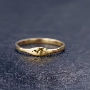 Puede incluir: Un anillo de oro con una banda lisa y pulida que presenta una sección frontal ligeramente más ancha y aplanada. El centro de esta zona plana está grabado con el contorno pequeño y sencillo de un ave en vuelo, como una marca sutil.