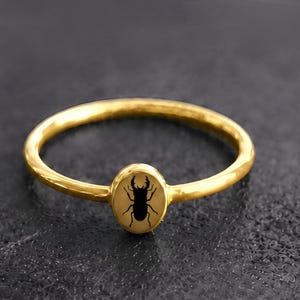 Può includere: Un anello color oro caratterizzato da una fascia sottile e liscia e da un piccolo elemento centrale di forma ovale. Il centro dell'ovale presenta una silhouette nera dettagliata di un cervo volante con mandibole prominenti.