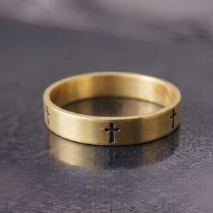 Pode incluir: Um anel de banda em tom dourado com acabamento em metal escovado e uma série de pequenos símbolos de cruz pretos, espaçados uniformemente, gravados ao redor da circunferência externa, criando um padrão repetitivo em toda a superfície.
