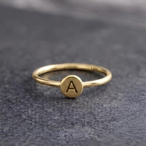 May include: A gold ring with a circular design featuring the letter "A" in the center. The ring is made of gold and has a thin band. The ring is set on a dark gray surface.