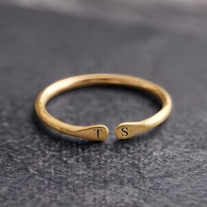 Peut inclure: Une bague ouverte de couleur dorée dotée d'un anneau lisse et arrondi. Les deux extrémités de la bague sont aplaties et présentent chacune une lettre gravée, avec la lettre T d'un côté et la lettre S de l'autre, créant un design personnalisé.