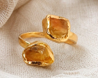 May include: A gold-toned open ring featuring two raw, amber-colored stones set in textured metal bezels. The band is smooth and circular, connecting the two irregular, faceted stones that sit at opposite ends of the open design, creating a bypass style.