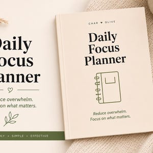 May include: A cream-colored Daily Focus Planner book with black text that reads "CHAR OLIVE", "Daily Focus Planner", and "Reduce overwhelm. Focus on what matters." A green pen rests on a textured beige fabric next to the planner.