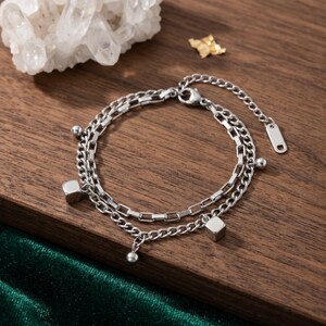 May include: A double-layered silver-toned metal chain bracelet featuring two distinct chain styles. The bracelet is adorned with small spherical beads and two polished cube-shaped charms hanging from the links, finished with a lobster clasp.