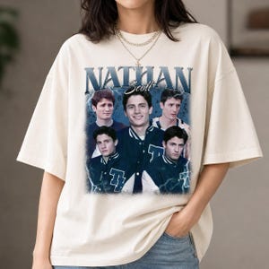 Puede incluir: Una camiseta de manga corta color hueso con un estampado gr&aacute;fico de Nathan Scott. El dise&ntilde;o incluye el texto NATHAN Scott en una fuente negrita sobre varias im&aacute;genes de la misma persona con una chaqueta universitaria sobre un fondo de rayos azules.