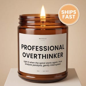 May include: A lit candle in an amber glass jar with a white label. The label reads MAVELLE, PROFESSIONAL OVERTHINKER, Light it when the spiral starts again. Pure analysis paralysis, gently interrupted, 9oz, SOY WAX. The candle has a single flame.