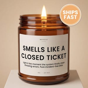 May include: A lit candle in an amber glass jar with a white label featuring the text MAVELLE, SMELLS LIKE A CLOSED TICKET, and Light it the moment the system finally stops throwing errors. Pure incident-free bliss. 9oz soy wax is printed at the bottom.