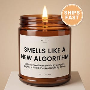 May include: A lit candle in an amber glass jar with a white label that reads MAVELLE, SMELLS LIKE A NEW ALGORITHM, Light it when the model finally compiles. Elegant solution energy, beautifully solved. 9OZ . SOY WAX. The candle has a single flame.