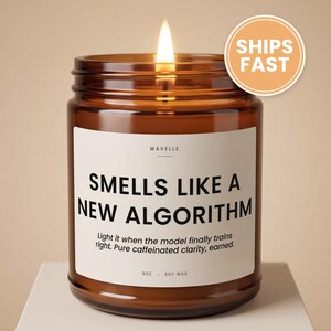 May include: A lit candle in an amber glass jar with a white label that reads MAVELLE, SMELLS LIKE A NEW ALGORITHM, Light it when the model finally trains right. Pure caffeinated clarity, earned., 9OZ, SOY WAX. The candle has a single burning wick.