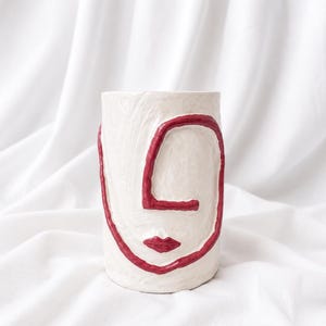 Pode incluir: Um vaso de cerâmica branco com um contorno vermelho de um rosto. O design inclui uma linha contínua para o olho e o nariz, e um pequeno par de lábios. O vaso é cilíndrico e está sobre um fundo de tecido branco.