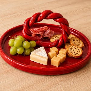 Pode incluir: Uma bandeja de servir em cerâmica vermelha brilhante com uma pega circular entrançada em relevo. A bandeja está disposta com um cacho de uvas verdes, fatias finas de salame, uma fatia de queijo brie, cubos de cheddar e pão torrado.
