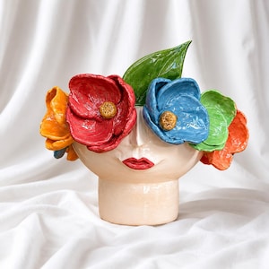 Pode incluir: Um vaso de cerâmica em forma de rosto, adornado com detalhes florais coloridos e grandes. As flores são em tons de vermelho, laranja, azul e verde, com uma folha verde. O vaso tem uma cor neutra, branco sujo.