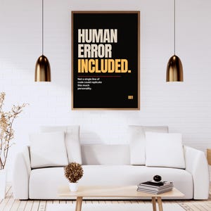 Op de afbeelding: Een rechthoekige wandprint in een dunne bruine lijst met een zwarte achtergrond en de tekst HUMAN ERROR INCLUDED in grote beige en gele letters, gevolgd door de kleinere tekst Not a single line of code could replicate this much personality en het getal 001.