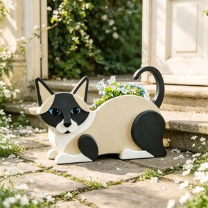 Op de afbeelding: Een katvormige plantenbak van hout met een crèmekleurig lichaam, zwarte oren, gezichtsmasker en staart. De plantenbak heeft blauwe ogen en een witte snuit, met een holle opening op de rug gevuld met kleine blauwe bloemen en groen.