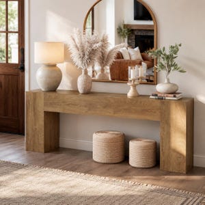 May include: A long, rectangular wooden console table with a light brown finish. On top sits a beige ceramic lamp, a textured vase with tall pampas grass, a wooden candle holder with two glass cylinders, and a small vase with green leaves.