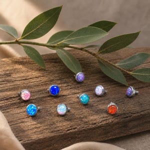May include: A collection of nine round, flat-back body jewelry studs featuring colorful synthetic opal cabochons in shades of pink, blue, turquoise, white, purple, and orange, each set in a metallic base with a small threaded post.