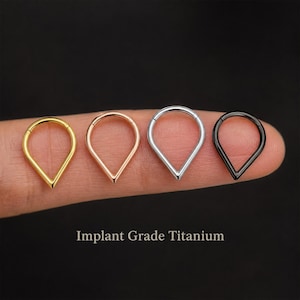 Puede incluir: Cuatro aros en forma de lágrima hechos de titanio de grado de implante. Los aros están dispuestos en fila y presentan acabados metálicos en oro, oro rosa, plata y negro. El texto Implant Grade Titanium aparece debajo de los aros.
