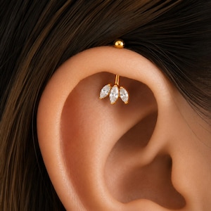 May include: A gold-toned curved barbell ear piercing featuring a decorative charm with three marquise-cut clear stones arranged in a fan-like cluster. The jewelry includes a small spherical ball end at the top and a secure post through the cartilage.
