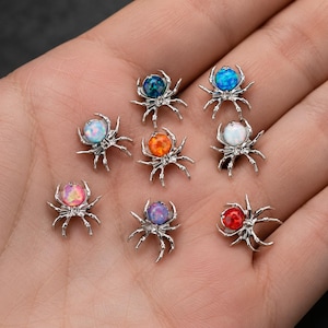 May include: A collection of eight silver-toned spider-shaped charms, each featuring a round, iridescent cabochon stone in various colours including blue, orange, white, pink, purple, and red, arranged in a cluster on a flat surface.