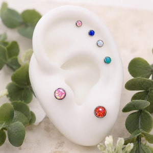 May include: A white ear model displaying six circular stud earrings with metallic settings. The studs feature various colored opal-style stones, including pink, blue, white, teal, and red, arranged in a vertical line and two lower positions.