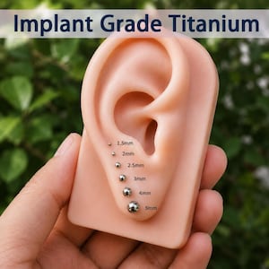 Puede incluir: Un modelo de oreja de silicona beige que muestra seis pendientes esféricos de color plateado en tamaños graduados de 1,5 mm a 5 mm. El texto Implant Grade Titanium es visible en la parte superior. Cada pendiente tiene su medida métrica en milímetros.