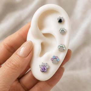 May include: A set of five silver-toned paw print stud earrings displayed on a white ear model. Each earring features a central heart-shaped gemstone in black, white, or purple, surrounded by small clear, blue, or pink accent stones.